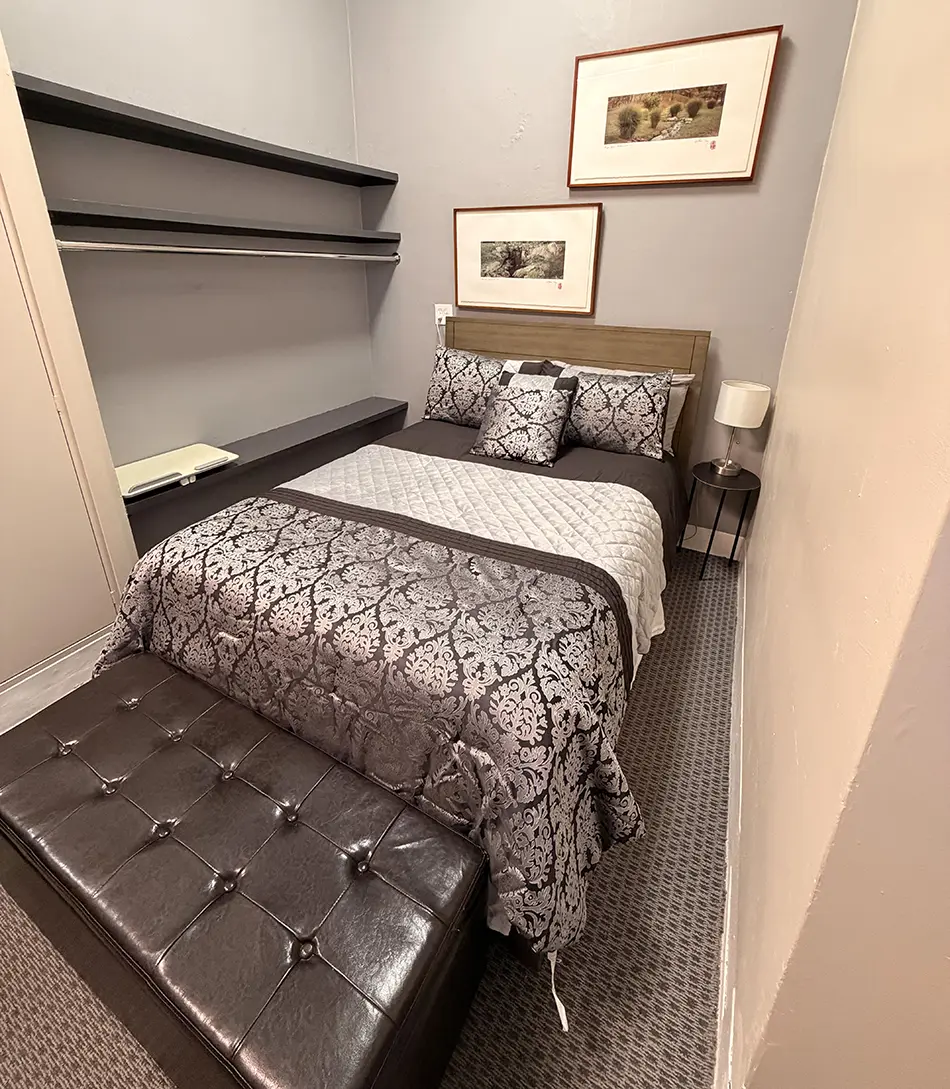 Queen bed with gray damask bedding and matching throw pillows, a dark tufted bench at the foot, and a small nightstand with lamp on the right; built-in shelves on the left and framed art on the wall above the headboard.