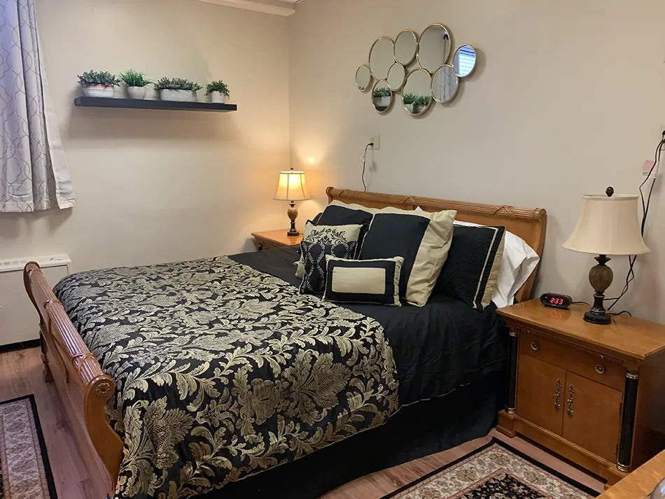 Cozy bedroom with a wooden bed, black and gold floral comforter, multiple decorative pillows, two nightstands with lamps, and circular mirror wall art with small plants above the shelf.