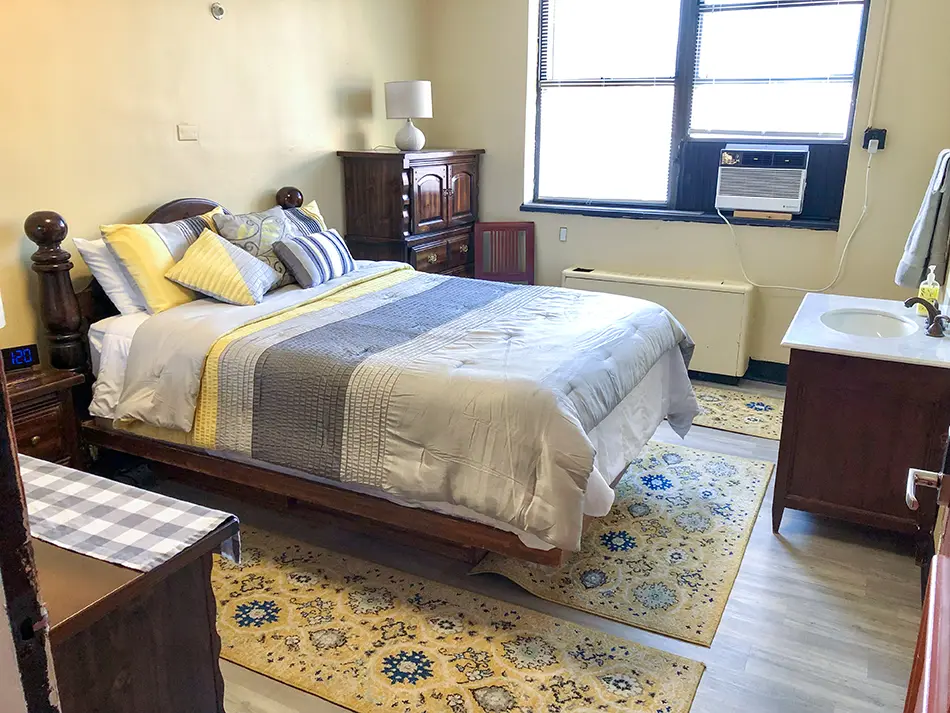 Hotel bedroom with a queen bed, yellow walls, and dark wood furniture; patterned rug, window with an air conditioner, and a sink vanity to the right.