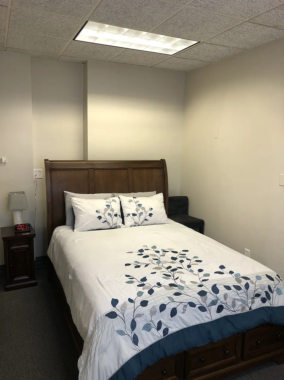 Bedroom with a wooden bed frame, white bedding with blue leaf pattern, two pillows, and a bedside table with lamp and digital clock reading 10:03.