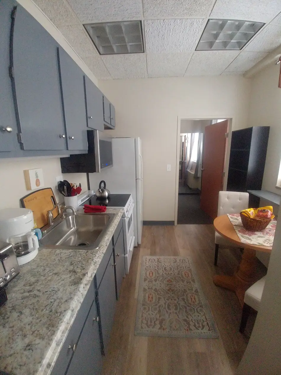 Compact kitchen with blue cabinets, white appliances, stainless sink, and a small dining area with a round table and a basket of snacks on the right.