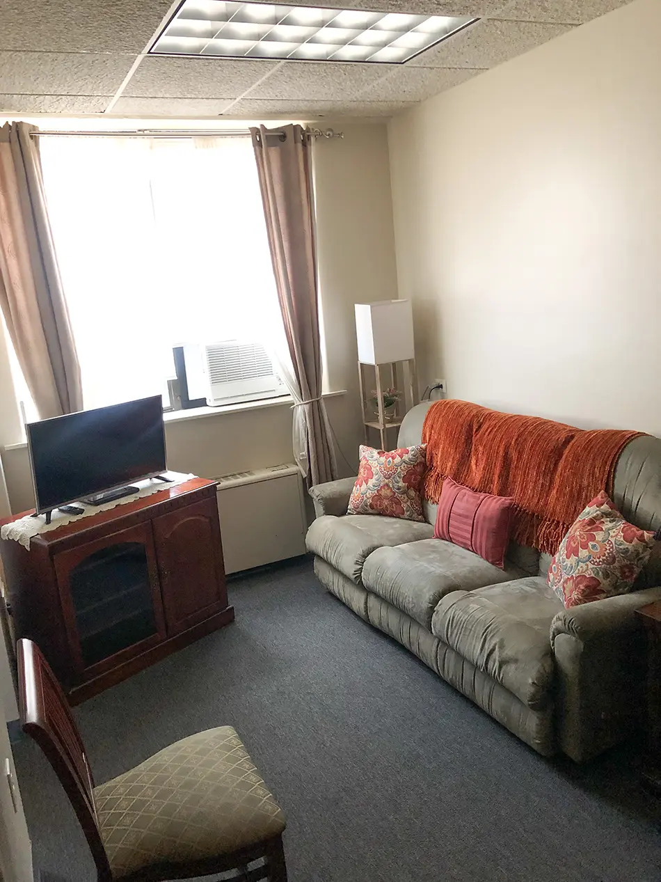 Small living room with a gray sofa, orange throw, floral cushions, and a TV on a wooden stand by a window with beige curtains and an AC unit in the window.