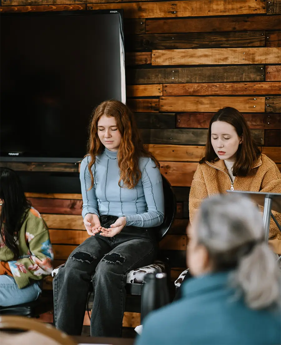 Two young women sit with eyes closed in a wood-paneled room during a quiet group meditation.